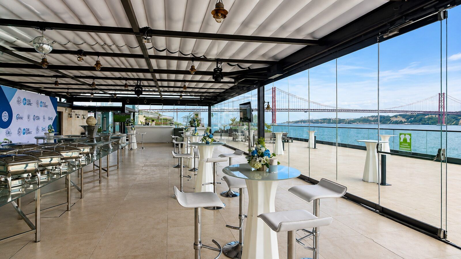 Riverside terrace set up with catering stations and cocktail tables overlooking the 25 de Abril Bridge in Lisbon