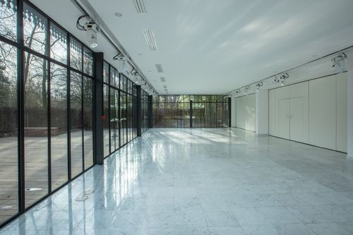 Empty glass-walled hall with marble flooring and floor-to-ceiling windows overlooking a garden