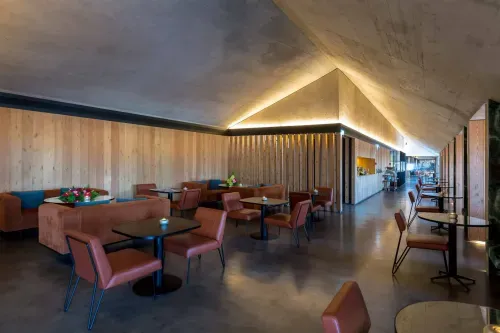 Contemporary indoor lounge with angled wood-panelled ceiling, warm accent lighting, and terracotta leather armchairs around dark round tables.