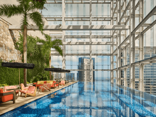 Rooftop sky pool in Singapore framed by glass beams and palm trees, with orange lounge seating and city skyscrapers visible through the glass facade.