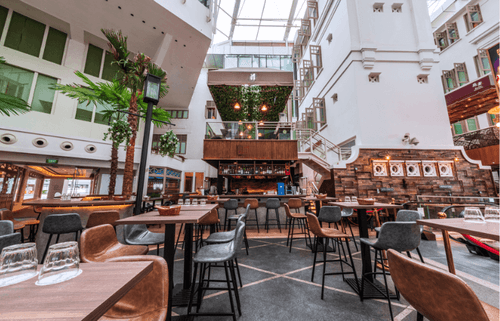 Sunken atrium courtyard with a central bar, wooden dining tables, palm trees and colonial-style architecture under a glass roof.