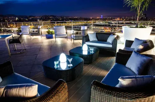 Rooftop lounge with rattan sofas, illuminated globe lights and cocktail tables at night overlooking the city skyline