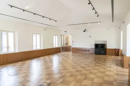 Empty conference hall in Milan with checkered parquet flooring, white walls with wood panelling, track lighting and tall windows.