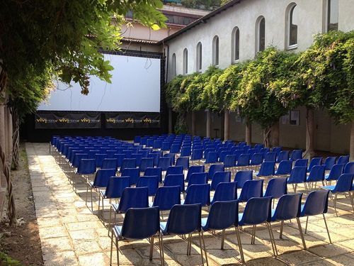 Renaissance courtyard in Milan set up as an outdoor cinema, with rows of blue chairs facing a large projector screen, framed by wisteria vines and historic buildings.