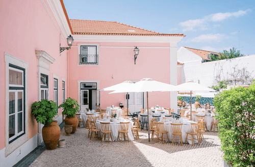 Cobblestone courtyard with round tables set under white umbrellas against a pastel pink Baroque facade with terracotta roof tiles and terracotta plant pots.