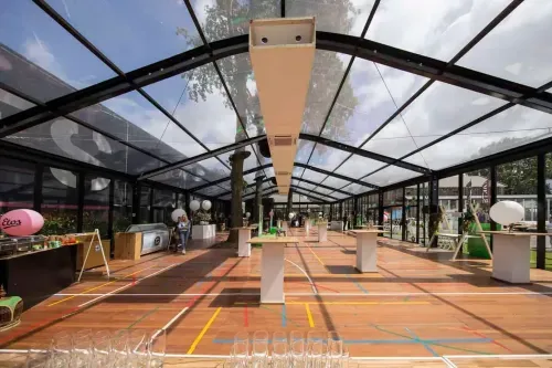 Glass-roofed pavilion with high-top event tables on a wooden floor, trees visible through the panelled walls