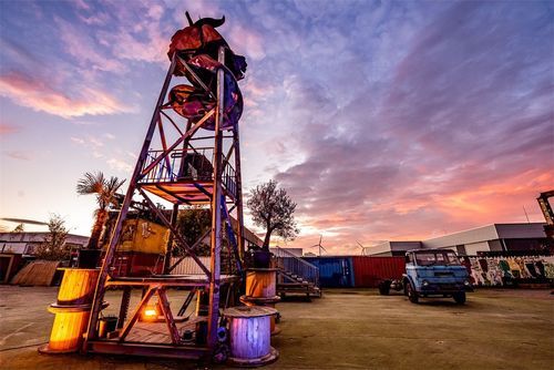 Open-air industrial site at sunset with a tall welded metal sculpture tower and a vintage truck