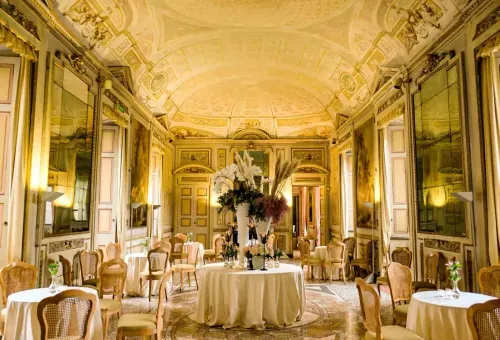 Neoclassical hall in Milan with gilded vaulted ceiling, painted wall panels and round dining tables with tall floral centrepieces, hosts up to 1,200 guests