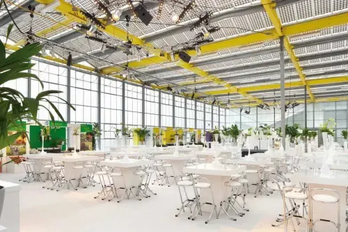 Large glass-roofed hall with rows of round tables, exposed yellow rigging and rows of windows