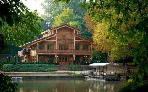 Wooden chalet on a lakeshore surrounded by trees, with a jetty and boats on the water
