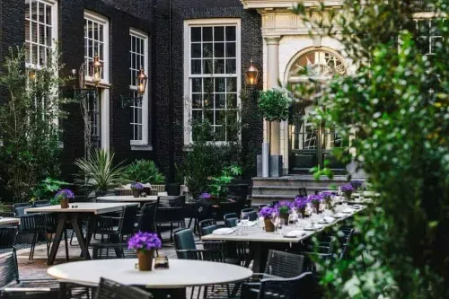 Courtyard garden set for dining with tables, purple flowers and greenery against a black-brick period facade