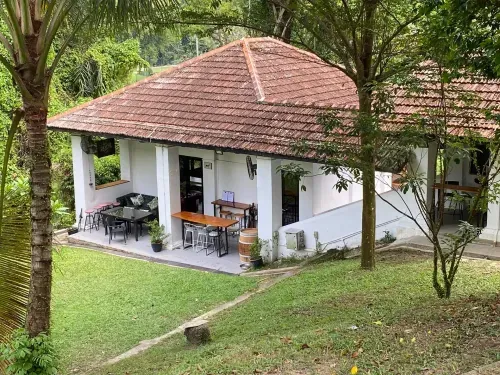 Colonial-style bungalow with a terracotta tiled roof, verandah seating and wooden tables, surrounded by tropical garden greenery.