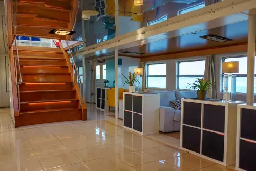 Interior of a double-deck event boat in Lisbon with wooden staircase, tiled flooring and lounge seating, river view through windows, hosts up to 230 guests