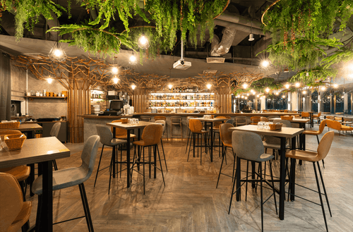 Indoor bar and dining area with hanging greenery, string lights and tree-branch decor, wooden high tables and mixed seating.