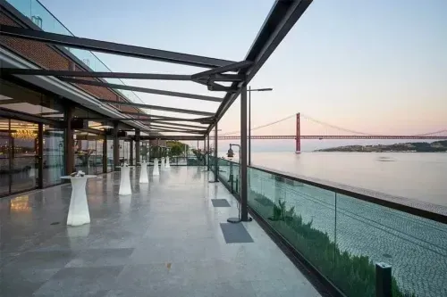 Covered glass-roofed terrace along the Tagus river in Lisbon at dusk, with high cocktail tables and marble flooring, the 25 de Abril bridge visible in the background.