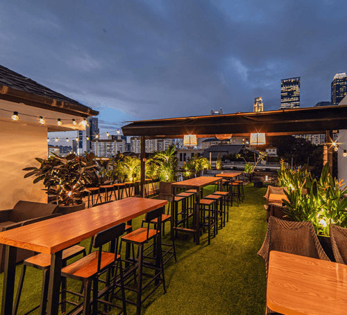Rooftop bar terrace in Singapore at dusk with wooden high tables, string lights, greenery and city skyline in the background.