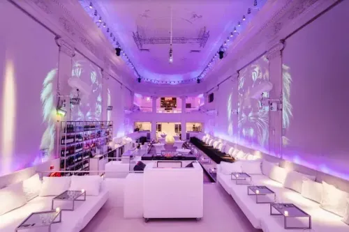 Neoclassical lounge venue in Amsterdam lit purple with white sofa seating, projected visuals and a stocked bar wall, hosts up to 450 guests