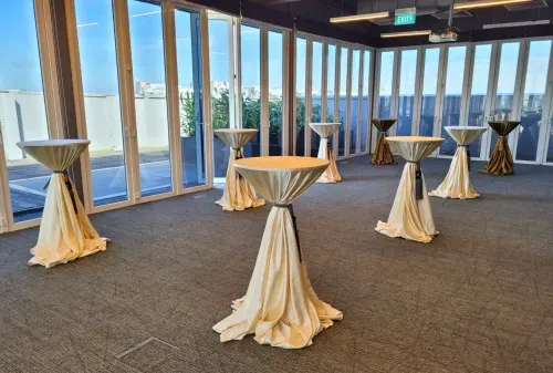 Rooftop event space with floor-to-ceiling glass walls, tall draped cocktail tables, and a view of the city skyline.