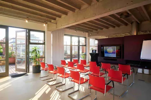 Sunlit loft workshop space in Amsterdam Overamstel with exposed concrete beams, red cantilever chairs and terrace access, hosts up to 100 people, 205 square metres
